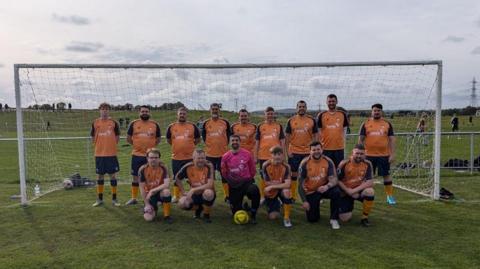 16 men on a green football pitch in front of a white net. 15 Men are wearing orange and black t-shirts and shorts while there is a man, the goal keeper, wearing a long sleeve pink shirt and black jogging bottoms.