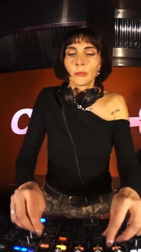 A woman, with short black hear wearing a black one-shouldered top, is DJing. Her hands are about to press the decks.