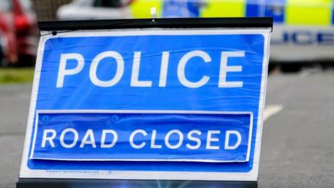 A police road-closed sign with a police vehicle behind it, slightly blurred. The road sign is blue with white writing.