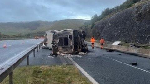 A grainy image of the M6 northbound around junction 36 for Kirkby Lonsdale. There is a burnt out lorry on its side in the middle of a carriageway. There are two workers wearing orange who stand by the truck. It is raining, with grey clouds hanging over the horizon. The road surface is wet.