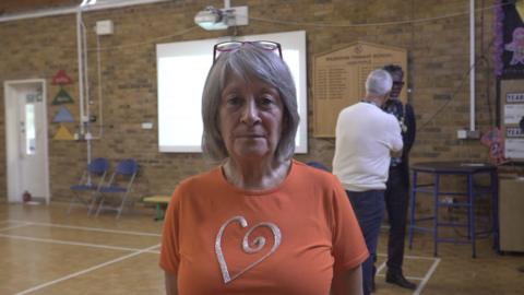 A woman hair with grey hair and wearing an orange t-shirt with a silver heart motif. She is standing in a school hall and looking at the camera. Two men stand behind her in the background talking to each other.