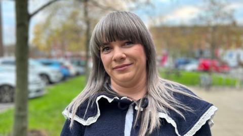 A woman with grey hair is standing outside. She is wearing a navy top. The background is out of focus, but trees, grass and a variety of parked cars can be seen.
