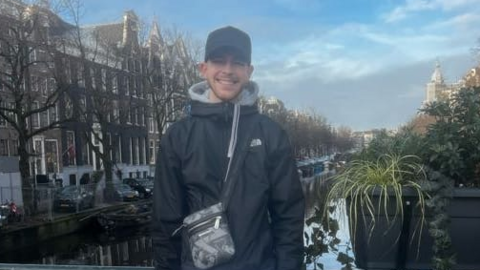 Robert Tyler-Jones standing on a bridge, in front of a canal and buildings, smiling at the camera, wearing a black cap, black top, trousers and a bag across him. He is standing next to a plant display.
