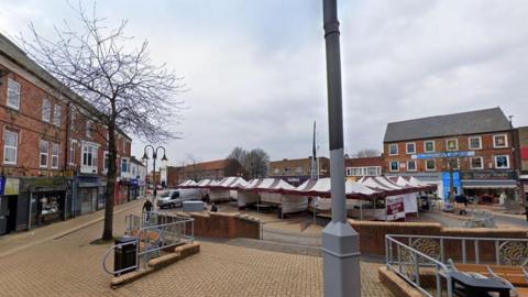 A view of the market place in Sutton in Ashfield. It is a light brown brick pedestrianised square with shops sitting on either side of temporary market stall tents.