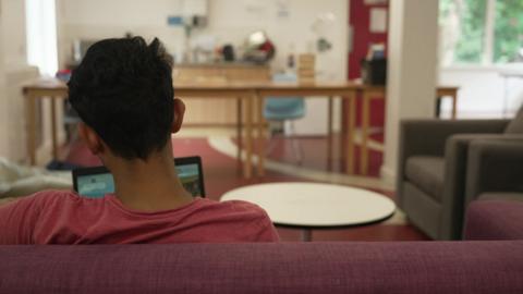 A resident of the hub sits on a laptop of the main communal area. He has short dark hair and is wearing a pink top and he is sat on a sofa. His back is facing the camera so all you can see of him is the back of his head, neck and shoulders.