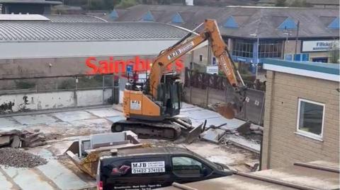 A building site with two vehciles parked alongside a skip and the remains of a demolished building