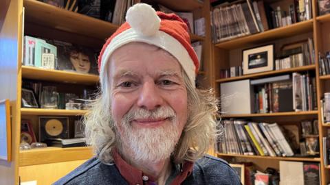 A man with white shoulder-length hair and beard and wearing a Christmas hat, a red shirt and a blue top smiles into the camera. Behind him are light brown wooden shelves full of records and trophy discs.