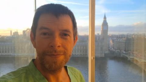 A man with a stubbly beard and short brown hair is standing at a window overlooking the River Thames and the Houses of Parliament.