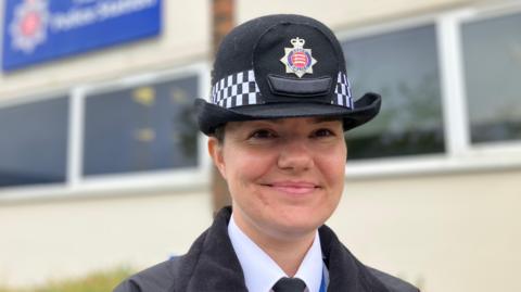 Chief Insp Emily Holmes is smiling in front of Harlow police station. She is wearing a black hat, black jacket, black tie and white shirt.