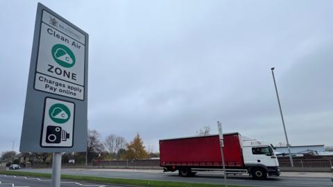 A road sign indicates Bradford's Clean Air Zone. It is captioned "Charges apply, pay online". A red lorry drives along the dual carriageway in the background.