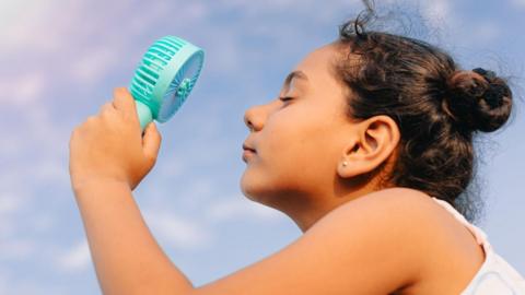 girl cools herself with portable fan with sun in sky