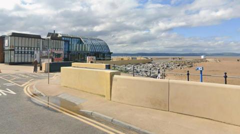 A view of the boathouse building at West Kirby beach with a yellow beach wall, sandy beach and a view of Welsh hills in the distance.