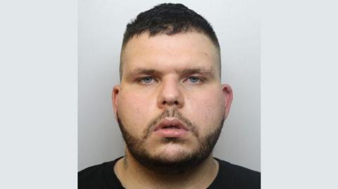 A police mugshot of Tom Kember. He is wearing a black t-shirt and standing against a grey background. He has dark brown hair, light blue eyes and and a dark beard. He is looking at the camera with a blank expression and his mouth slightly open.