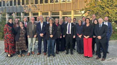 Men and women wearing suits and dresses and looking at the camera. They are all stood up. Below them is grey cobbled stone. Behind them is a grey building with glass windows. There are trees behind them on the right and a bush with pink flowers behind them on the left. 