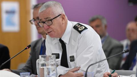 Jon Boutcher, he has white hair and glasses and is wearing a white shirt shirt with PSNI emblems. He is sitting at a table and is leaning forward speaking into a black microphone. A jug of water is in front of him.