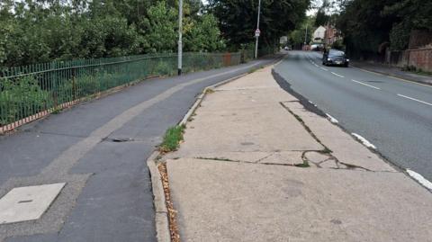 A layby next to a road, which disappears into the distance. There are cracks in the layby's concrete, as well as in the pavement on the left. A black car on the other side of the road is driving towards the camera. There are green trees on the far left and buildings in the distance.
