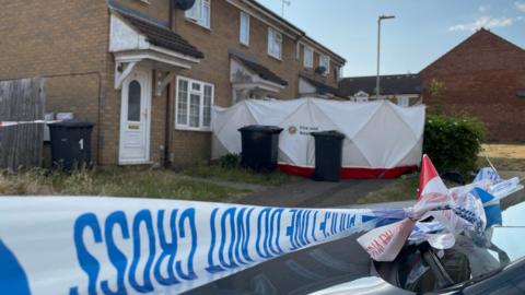 Blue and white police tape cordoning off the entrance to the house, which is mid-terrace and made of cream bricks. There is a white tent surrounding the entrance.