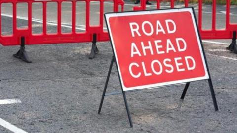 A red road sign reading "Road ahead closed" in the middle of a streey. There are red street barriers behind it.