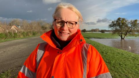 Jackie Hollowell, in a hi-vis jacket, stood by a rural road with flooded fields in the background