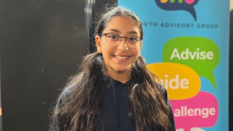 Ariyana is smiling as she stands in front of a pull-up banner which reads: Young Advisory Group, Guide, Advise Challenge. She has long dark hair in bunches and is wearing a par of glasses and a black hoodie.