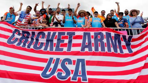 Fans, some wearing India shirts, stand and cheer above an enormous Stars and Stripes flag that has the phrase "Cricket Army USA" written on it