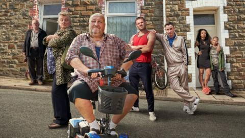 A group of people gathered in the street. They're in a V-formation behind one man, the central character, who is sitting in a mobility scooter.