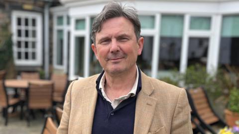 The President of the Scrutiny Management Committee, Deputy Andy Sloan stood wearing a beige blazer, blue jumper and a white shirt. He is stood outside in front of a conservatory smiling at the camera.