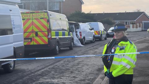 A police officer wearing a high vis jacket and bowler hat stands behind a blue and white strip of police cordon tape. Behind her is a residential street, with a number of police forensics vans along it.