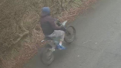 A person in a dark coloured coat, grey trousers and blue trainers riding a black bike along a road with grass and bushes in the background