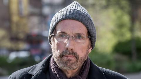 A man in his sixties wearing a grey bobble hat, dark winter jacket, a mauve scarf and glasses. He looks pensively just to one side of the viewer. He has a short beard and moustache.