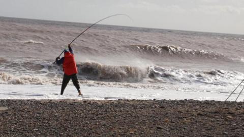 A man can be seen on a stony beach holding a fishing rod above the surf of a grey-brown sea near the shoreline. He has his back to the camera and is wearing a red jacket.