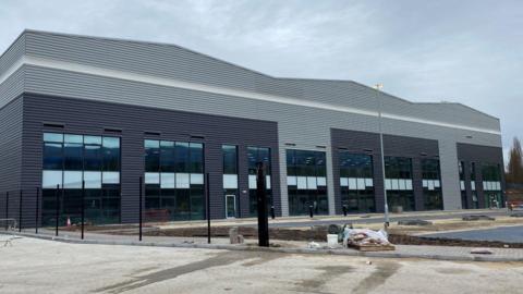 A grey warehouse with large windows along the front. The site looks empty, with a few items used in construction scattered around.