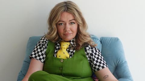 Ms Terry is sitting on a light blue chair. She is looking at the camera with a confused expression. She is wearing a black and white checked t-shirt, yellow tie and green waistcoat and trousers. She has long blonde hair. It is a PR promo image.