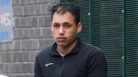 Man in a black zipped jacket stands outside by a brick wall and metal fencing, looking toward the camera.