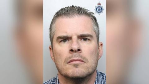 Mugshot of Simon Clark, 46, who has grey short hair and a grey beard. He frowns at the camera. 