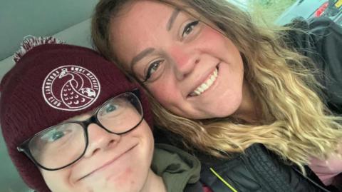 A selfie of Harry Basham with his mum Leanne Golding. Harry is wearing a burgundy hat with 'Taunton Town Football Club' written on it. He is also wearing glasses. Leanne Golding has long wavy blonde hair and is wearing a dark grey jacket with a pink top.
