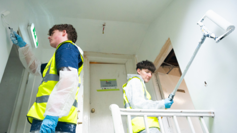 Two young people wearing hi-vis vests painting a hallway white. They are both holding paint rollers.