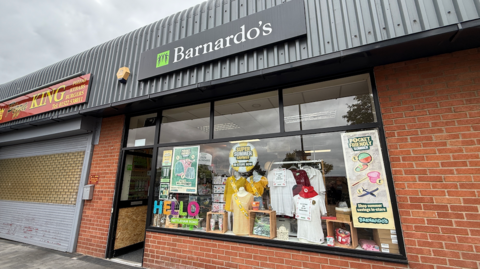 An outside view of a charity shop called Barnardo's which has part of its front door boarded up with a wooden panel. Clothes and signs are in the shop window. 