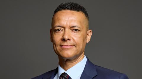 Clive Lewis looking at the camera wearing a blue jacket, a light blue shirt and a dark blue tie. He's looking at the camera with a slight smile on his face