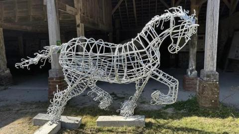 A sculpture of a running horse made out of steel. The metal is a light silver colour and the sculpture is not solid, with the main body made out of mostly circular pieces of metal with rods attaching them together. Three of its four hooves are bent and in the air, making it appear as if it is trotting or cantering. The horse's tail has been made with curvy pieces of metal, creating a flowy effect. The sculpture is on a patch of grass in front of what appears to be a barn.