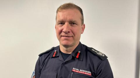 Wayne Bowcock is standing up wearing his RBFRS uniform, a blue shirt which has "Royal Berkshire Fire and Rescue Service" sewn into it.