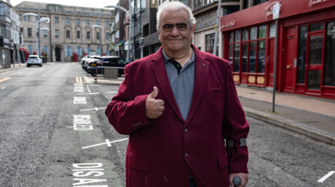 Campaigner Brian Roberts, with grey hair and wearing a maroon jacket, smiles and gives the "thumbs up" sign while standing next to new disabled parking bays
