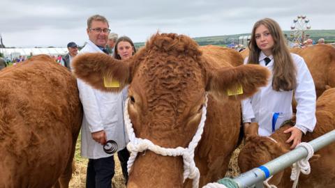 A young girl in a white jacket holds a brown calf gently around its neck, surrounded by other large brown cows. A man and another girl also in white jackets stand behind.