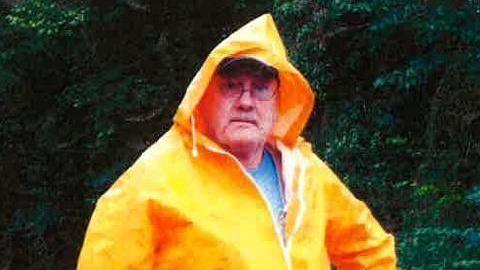 A man with a light blue t-shirt underneath a yellow/orange anorak which is zipped up with a hood on. He appears between 50 and 70 years old. He has glasses on and a neutral expression.