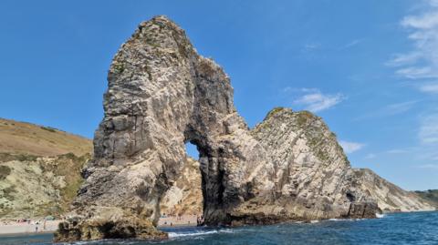 A large rock cliff arch sticks out into the sea. People can be seen on the beach through the arch. There are cliffs behind topped with browning grass. The sky is blue.