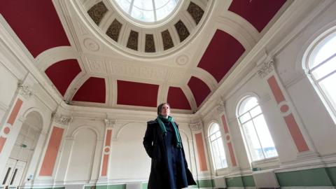A woman with short dark hair is wearing a navy blue winter coat and green scarf. She is stood in a derelict building which has an ornate ceiling with a glass dome and red paint. The walls are decorated with plaster and peach coloured paint