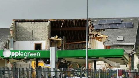 Aftermath of explosion at petrol station. A large hole can be seen in a nearby building and a digger is seen at the site.