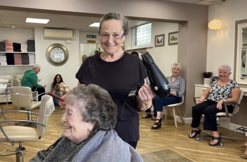 Sandra Handel on her last day at work at her salon in Highbridge. She is smiling at the camera and holding a hairdryer in one hand and a comb in the other. In the background are customers in chairs also smiling, and Sandra is standing over a customer with a grey towel wrapped around them