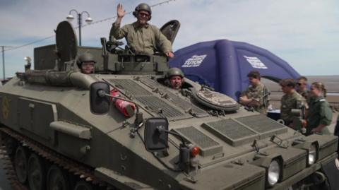 A green military tank is driving down a street. Two men wearing helmet and sunglasses are peering through an open hatch at the top of the tank, while another is doing the same just in front. Other members in green military uniforms are standing by the side looking as it drives past.