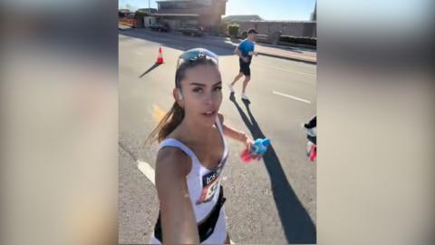 A woman with long brown hair wearing sports kit films herself while running along a road